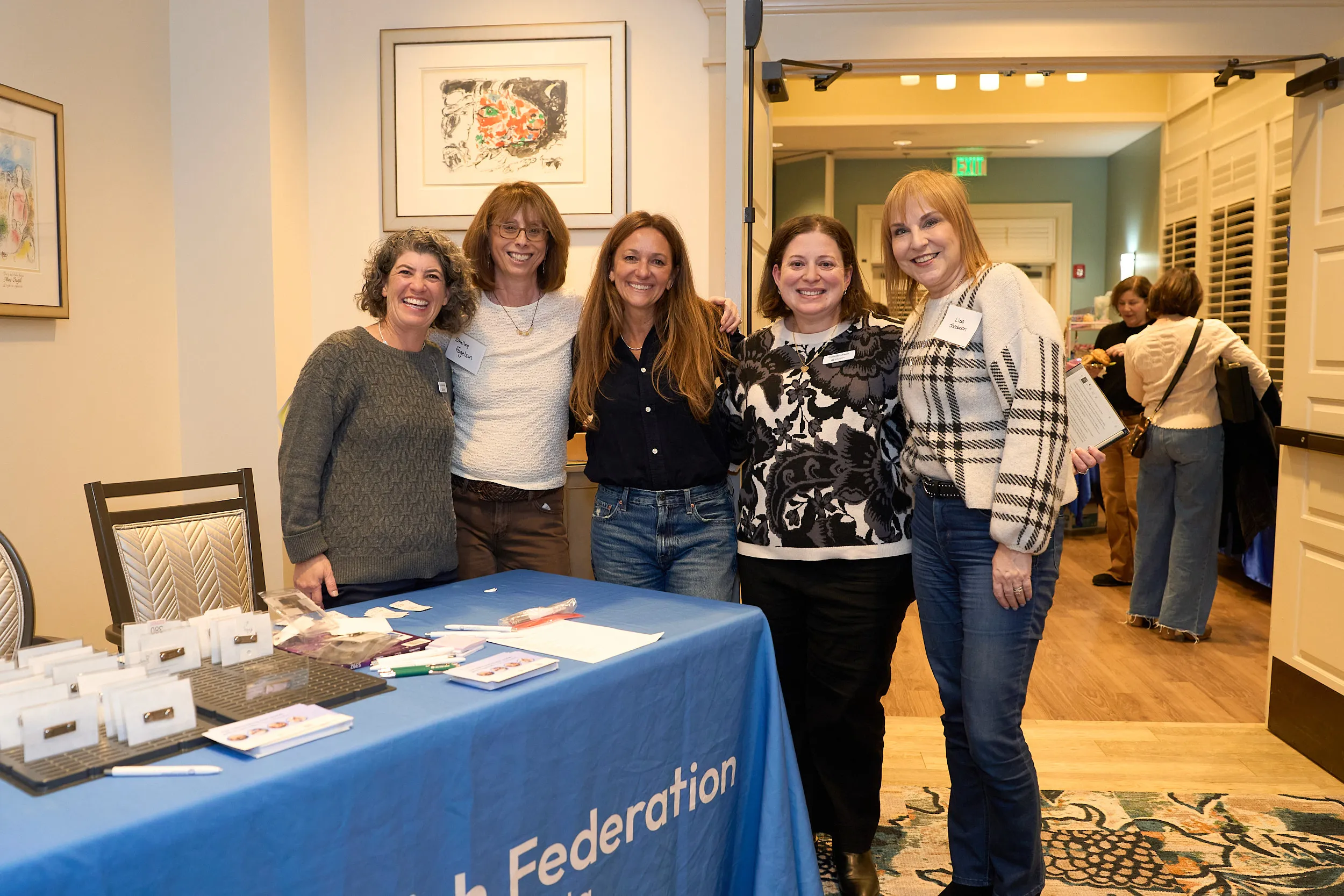 Five smiling women stand together behind the registration table at a Jewish Federation of Greater Atlanta event, welcoming guests as attendees arrive in the background.
