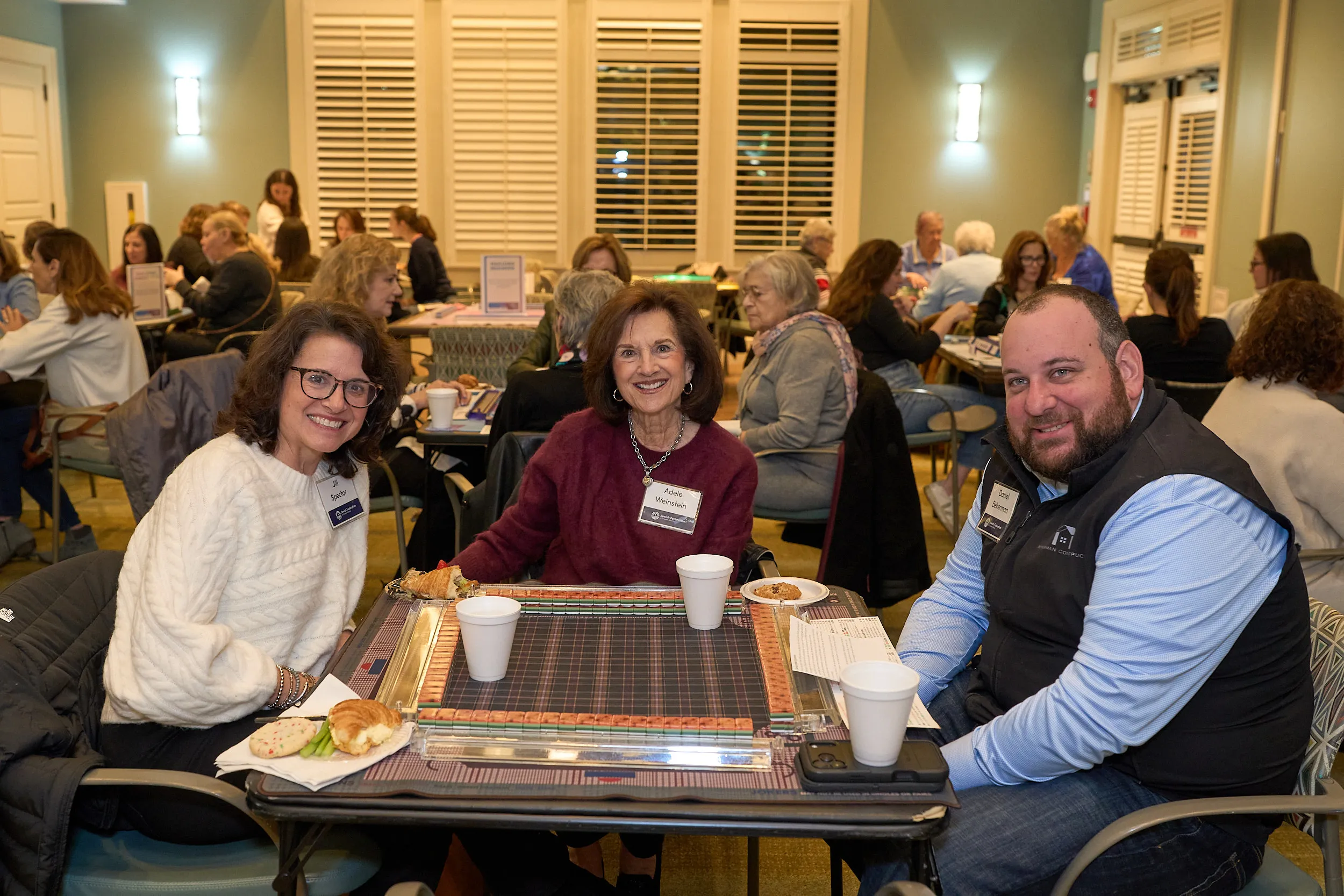 Three participants smile at the camera while seated around a game table during a Jewish Federation of Greater Atlanta event, with other attendees engaged in conversations throughout the room.