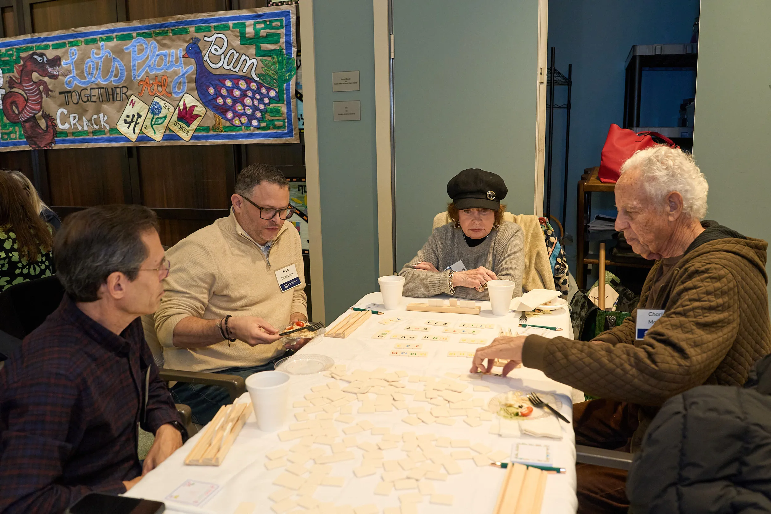 Four participants sit around a table playing a tile-based game during a Jewish Federation of Greater Atlanta community event, with colorful artwork displayed in the background.