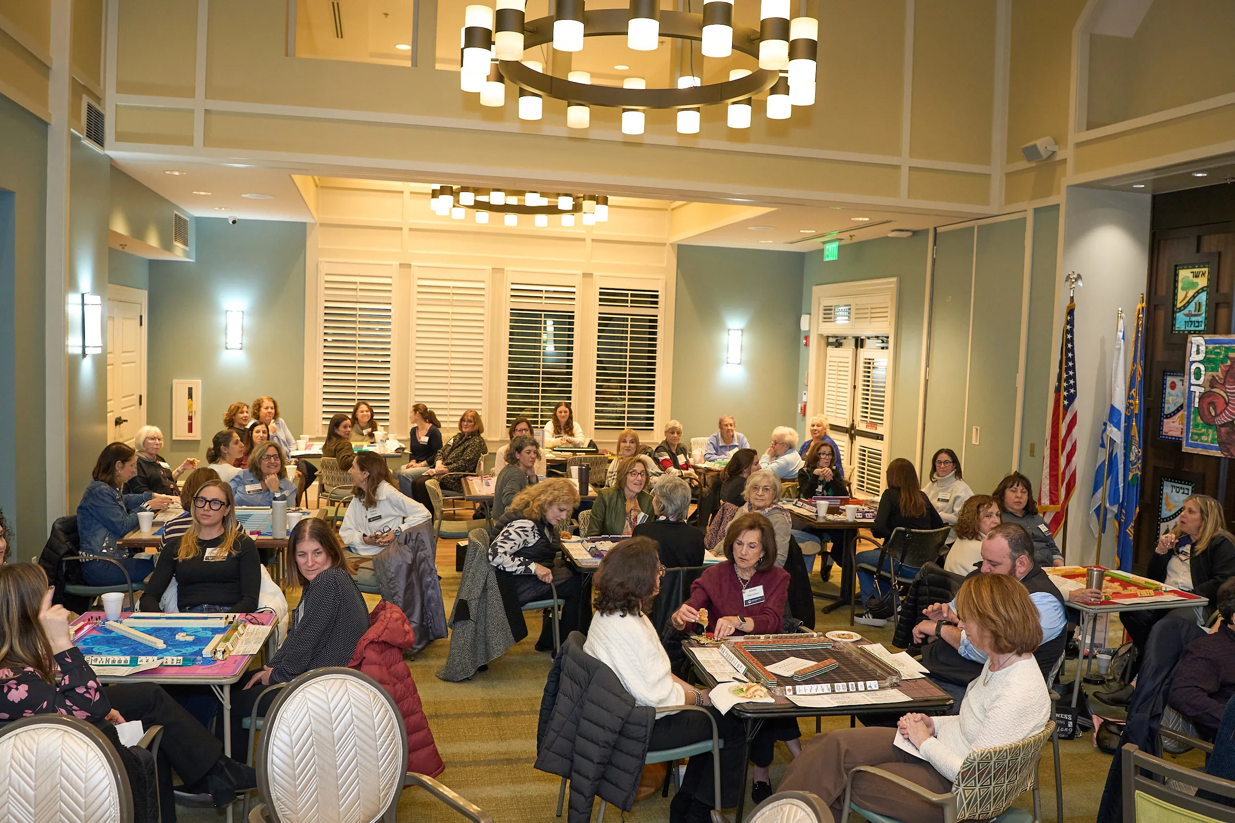 Attendees gather around multiple tables playing board and tile games during a Jewish Federation of Greater Atlanta community event in a bright meeting space.