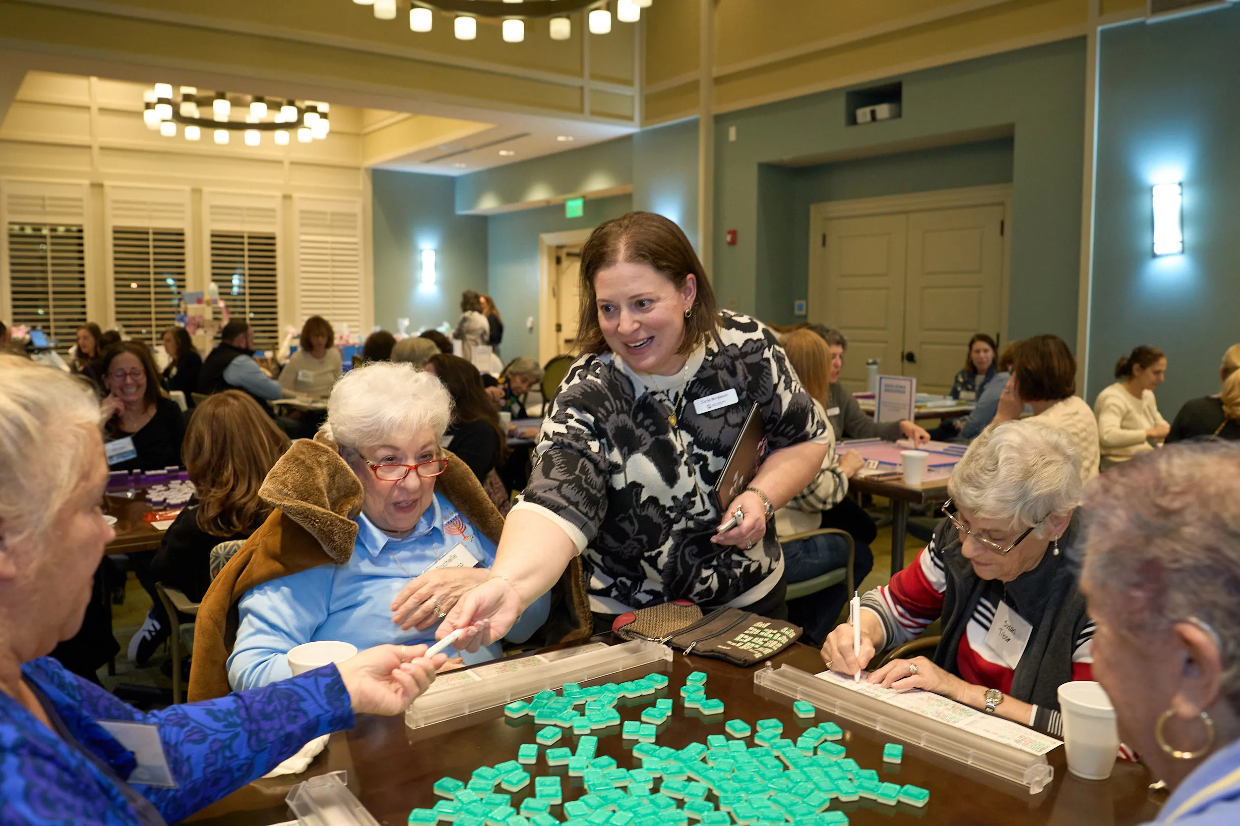Carla, JGather's program director, stands beside a table assisting participants during a tile game at a JGather community event, while attendees enjoy conversation and activities throughout the room.