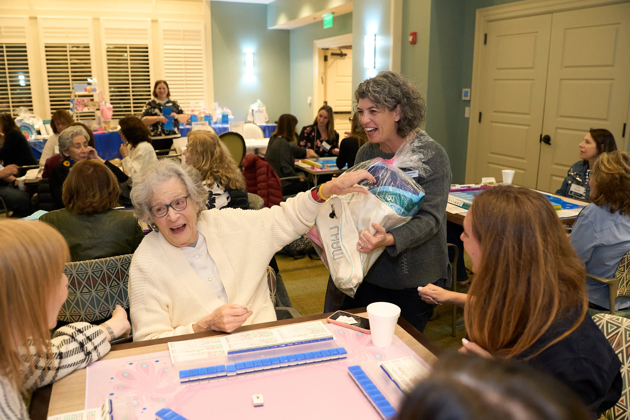 Laura Venti, JGather Connector, smiles as she presents a prize to a participant during a JGather game event, while attendees enjoy games and conversation around the room.