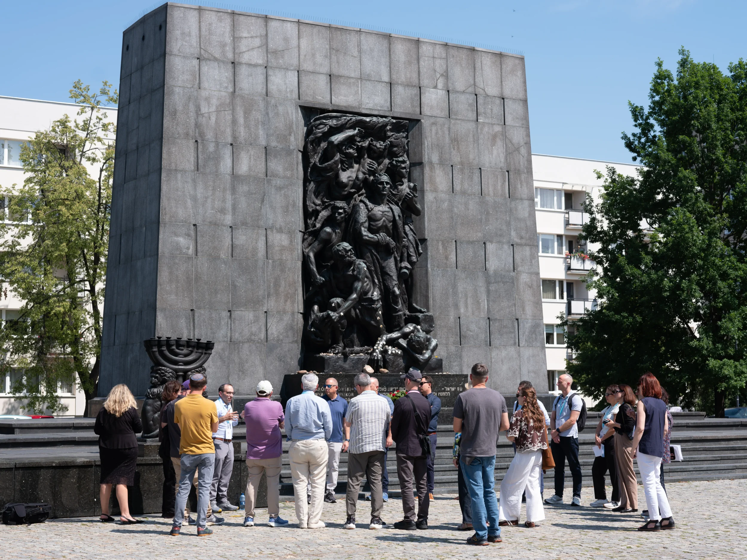 JDC study mission participants gather at a large Holocaust memorial featuring a sculpted relief and menorah in Warsaw, Poland.