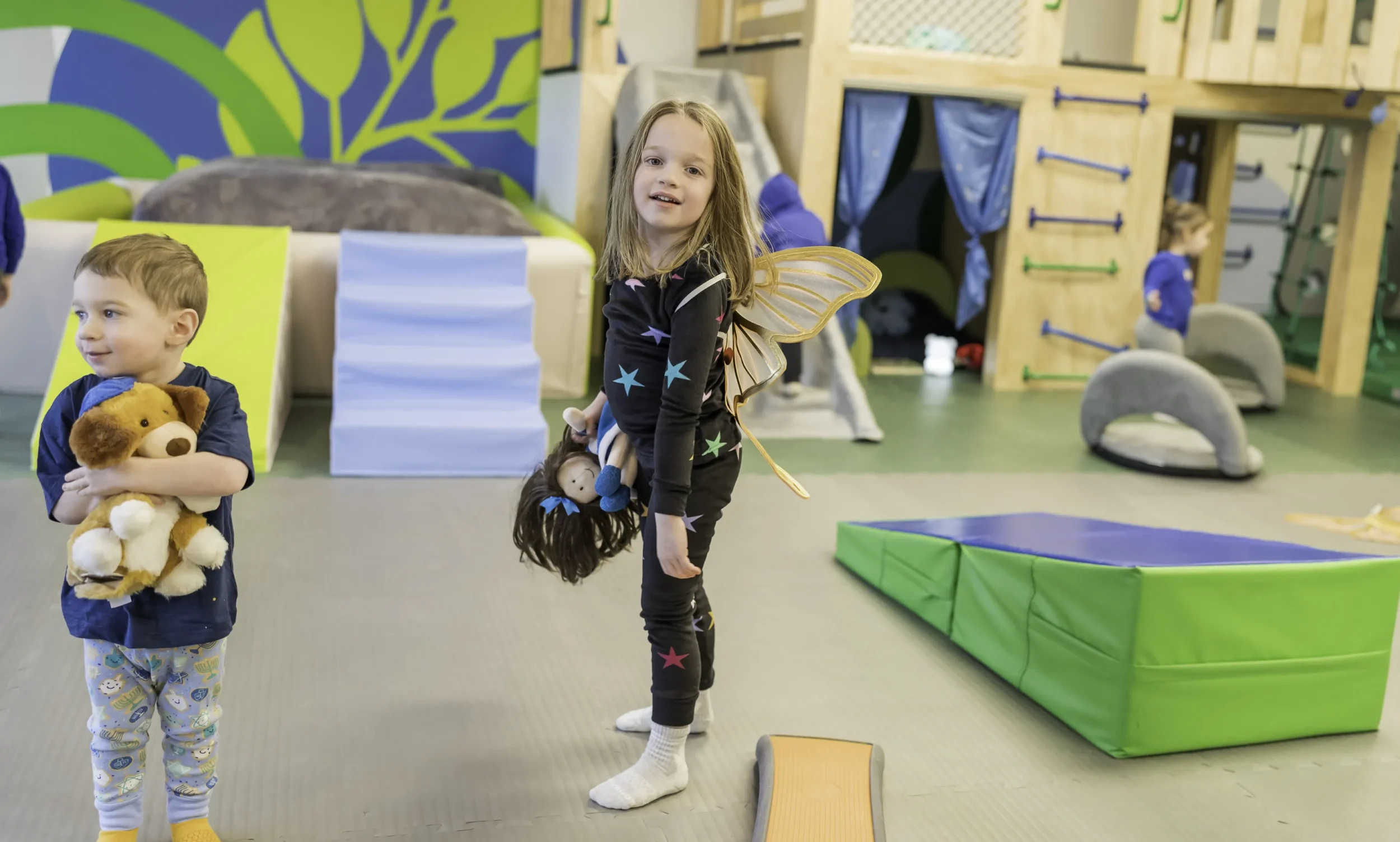 Little girl looks at camera, wearing butterfly wings. A boy in the corner holds a stuffed animal.