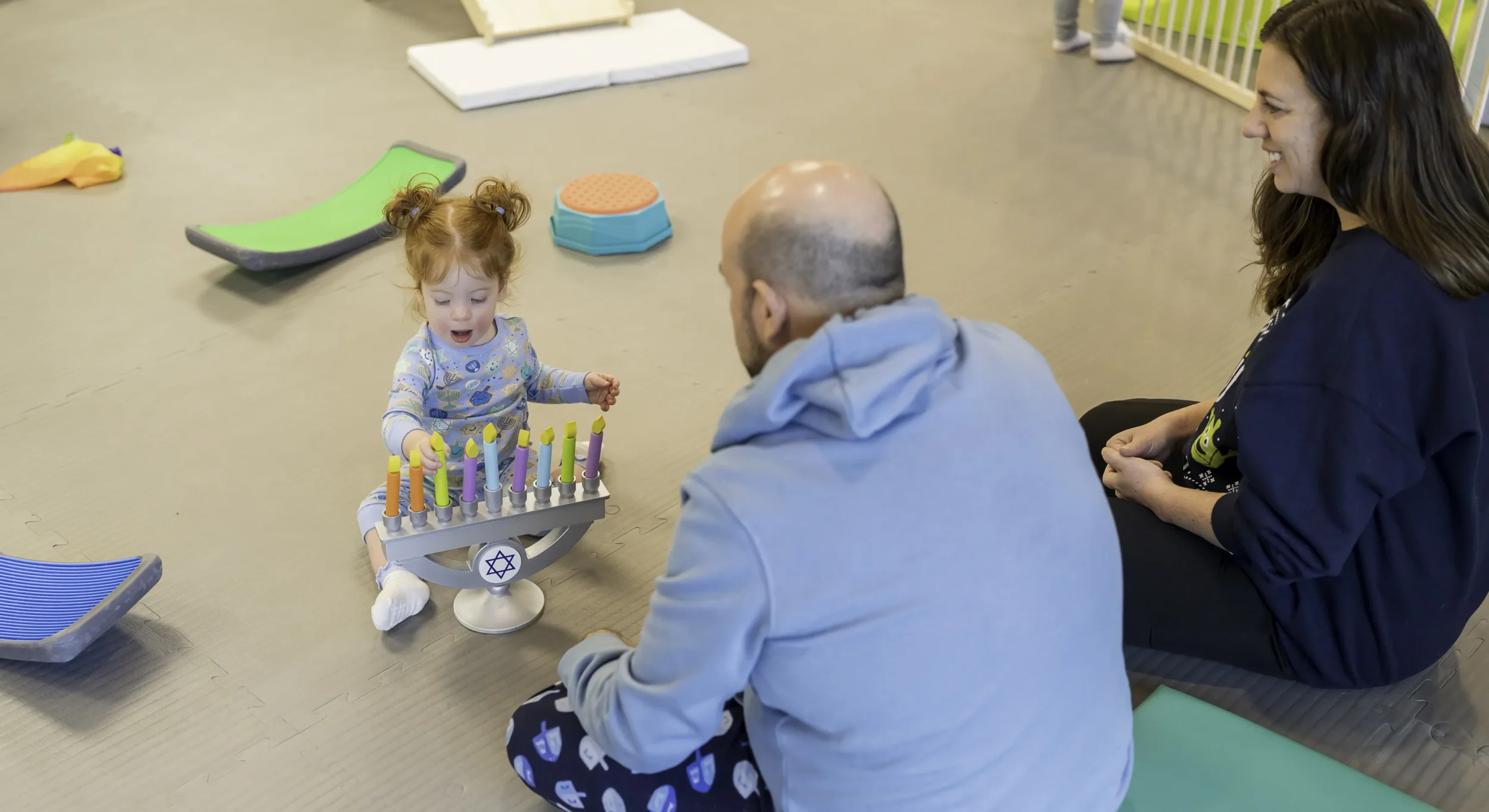 Mom and dad sitting with their daughter at PJ Library Hanukkah Party. Daughter plays with wooden candles.