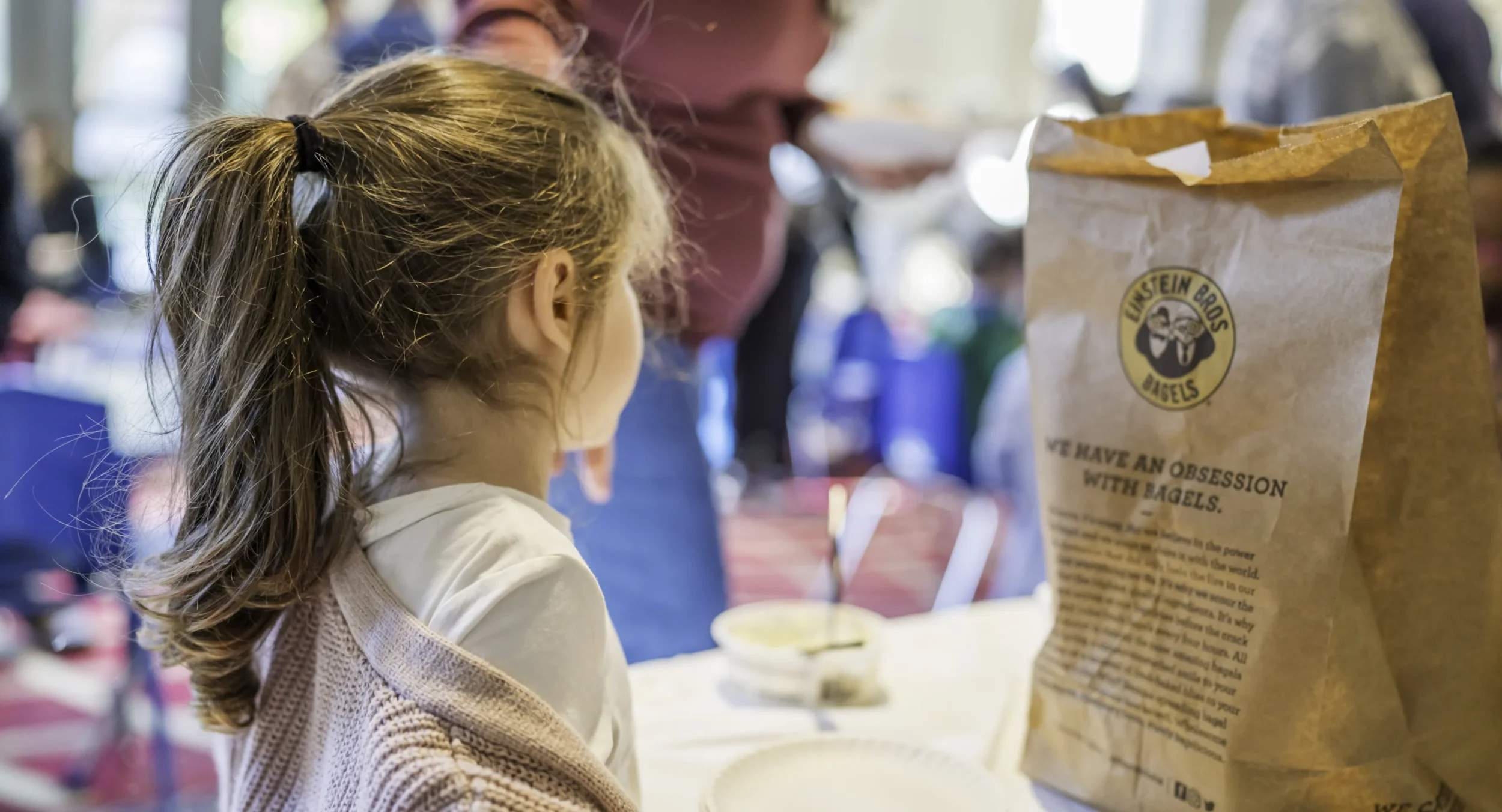 Little girl looks off into distance, a brown bag of Einstein Bros Bagels sitting on the table