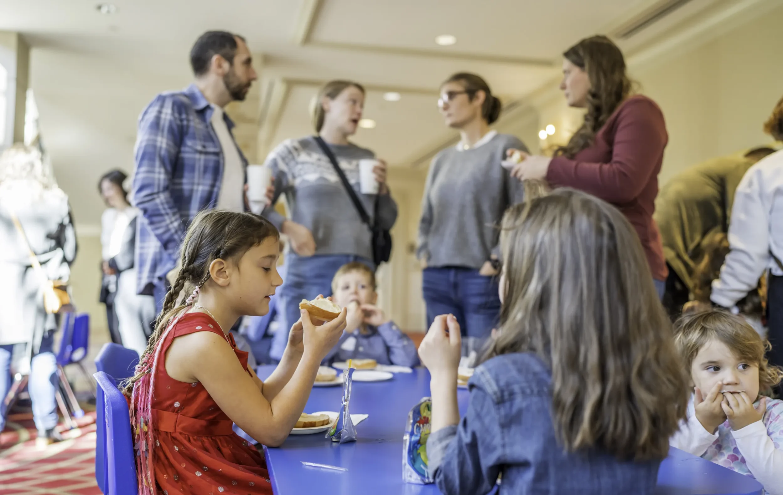 Adults standing in background talking, while kids eat food on table, at PJ Library event